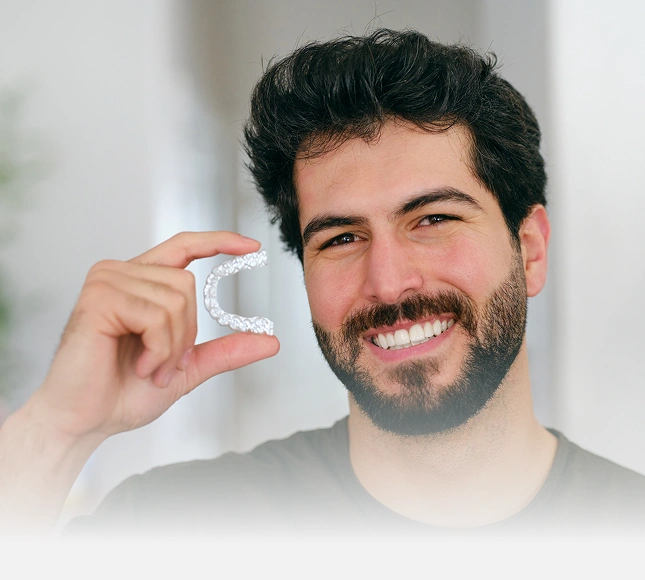 Man smiling while holding clear orthodontic aligner in front of his face indoors