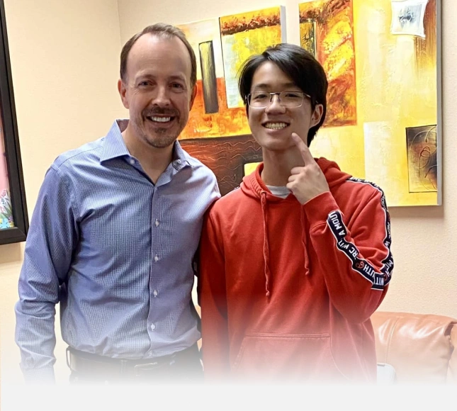 Two men smiling at camera in orthodontics office with artwork on walls behind them.