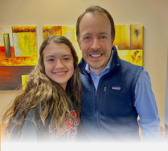 Two people smiling at camera in orthodontics office with colorful artwork on wall behind them.